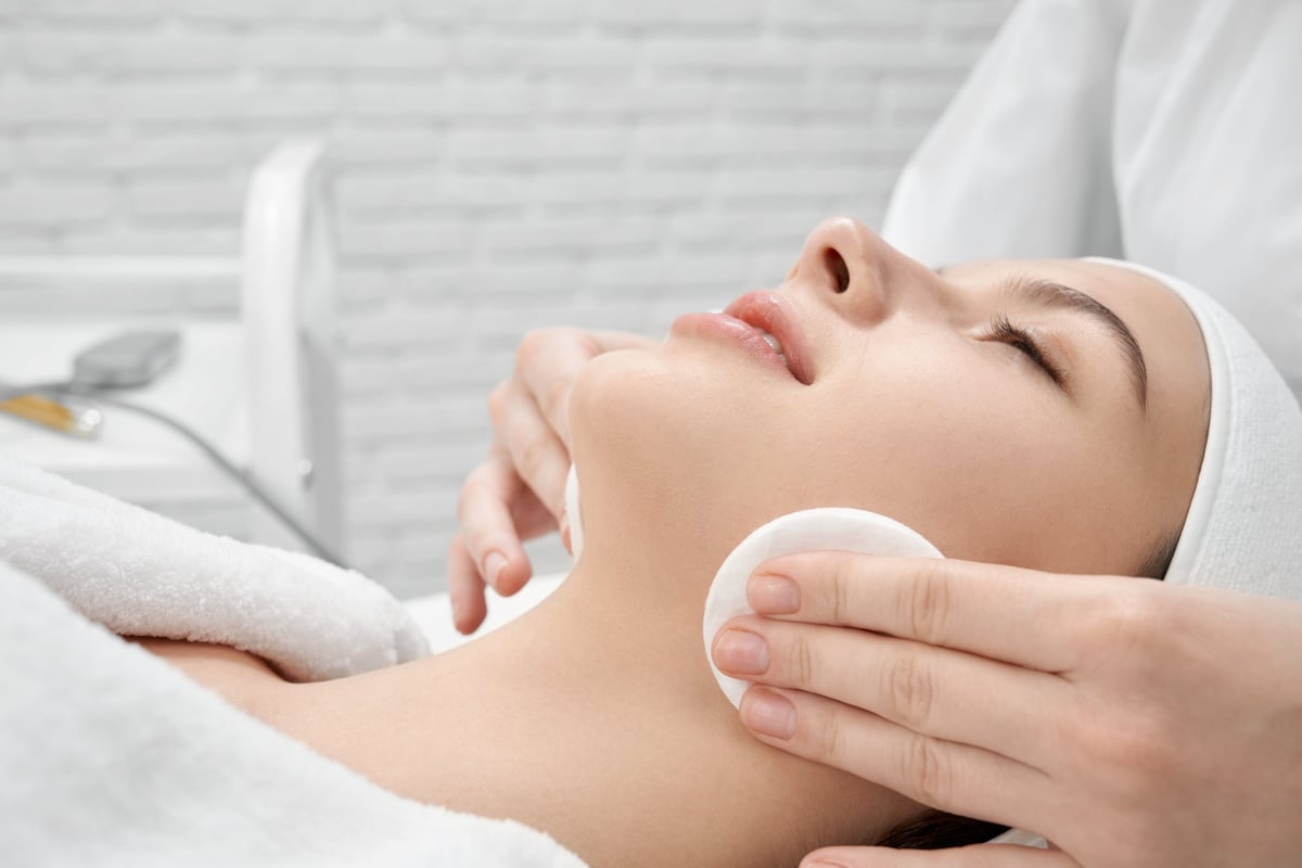 Young woman in white bathrobe lying with closed eyes in cosmetology cabinet. Professional beautician cleaning face of patient white cotton discs. Client enjoying.