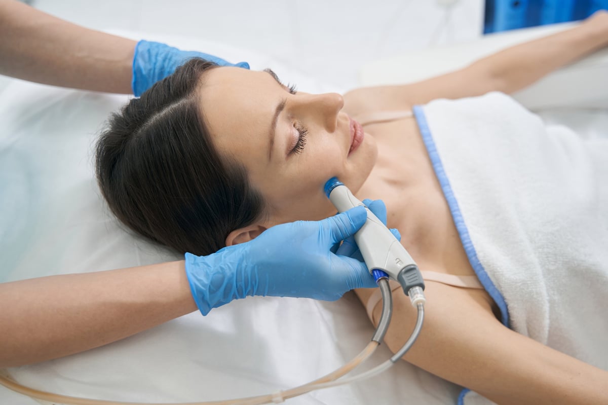 Cosmetologist holding Hydradermabrasian device and moving it along female cheek for improving wrinkles and fine lines