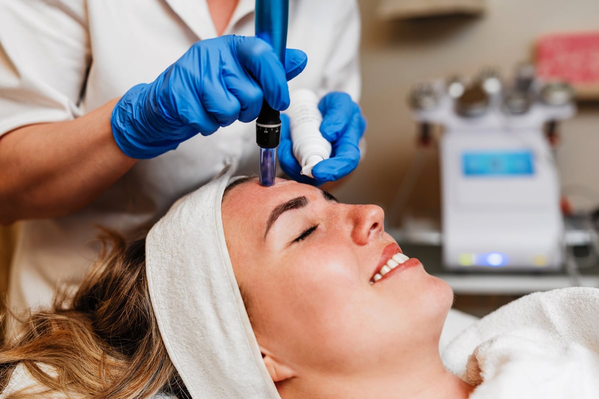 Esteticista aplicando suero con guantes azules durante tratamiento de microagujas dermapen en rostro de mujer joven