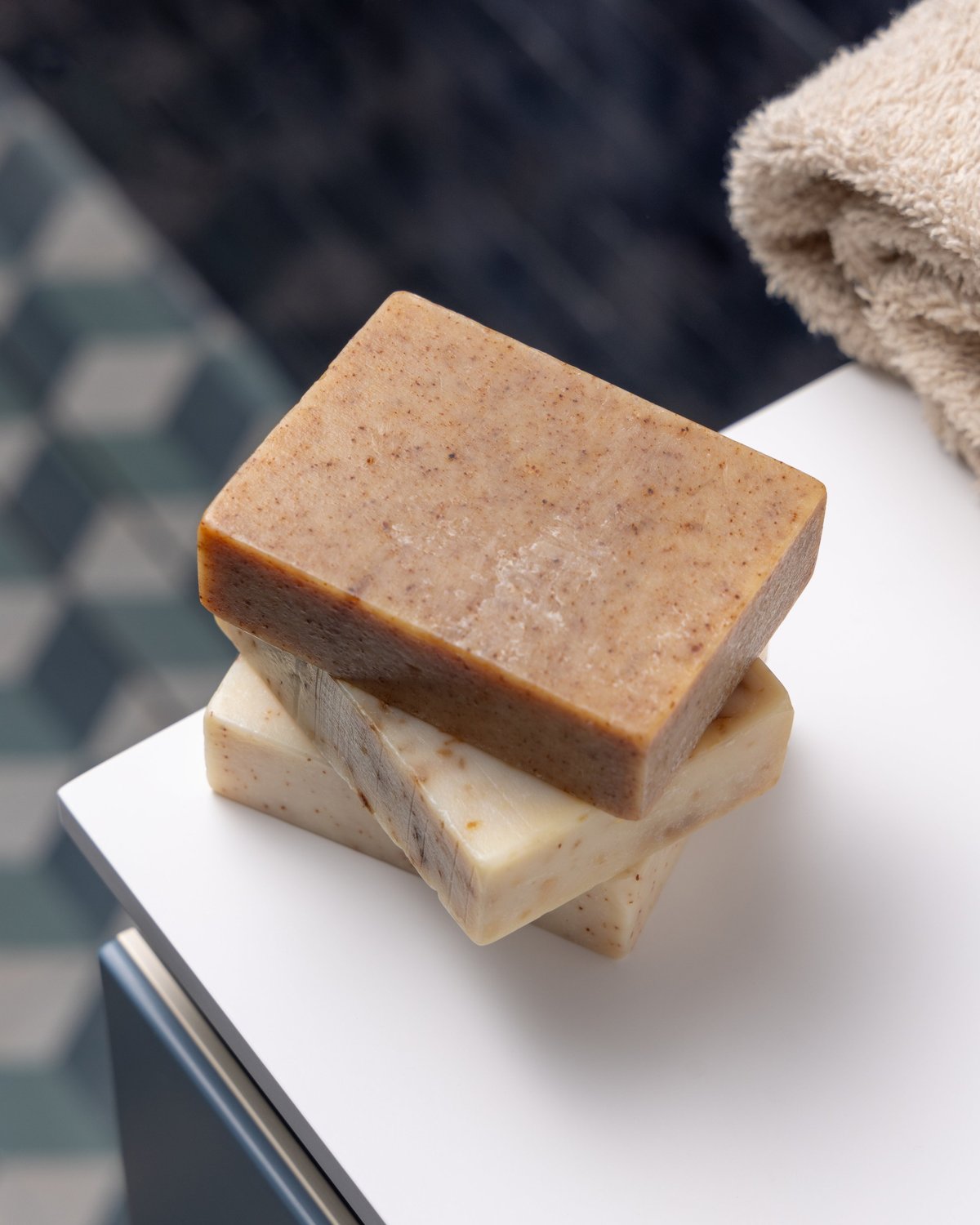 Brown and beige handmade solid soap bars near beige towel in blue tiled bathroom. Natural solid exfoliating scrub soap set for body cleansing, hygiene and skincare